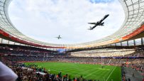 Two airplanes fly over a packed rugby stadium during a match, a large screen displaying Rugbys Greatest Rivalry