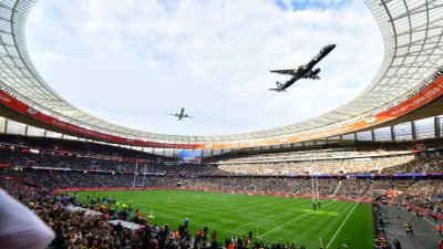 Two airplanes fly over a packed rugby stadium during a match, a large screen displaying Rugbys Greatest Rivalry