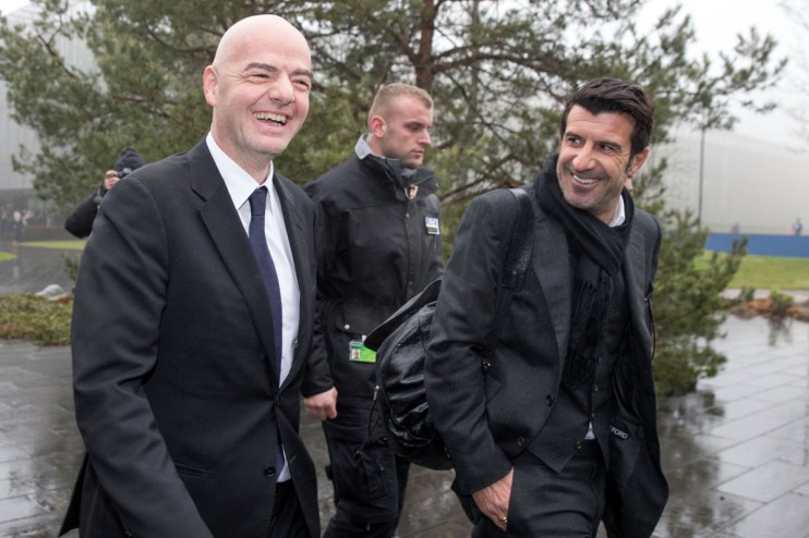 Gianni Infantino and Lu&iacute;s Figo smiling, walking outdoors with security, on a wet day.