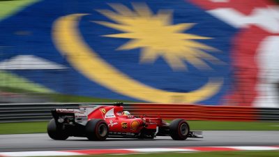 Red Ferrari F1 car speeding past a blurred Malaysian flag banner on a race track
