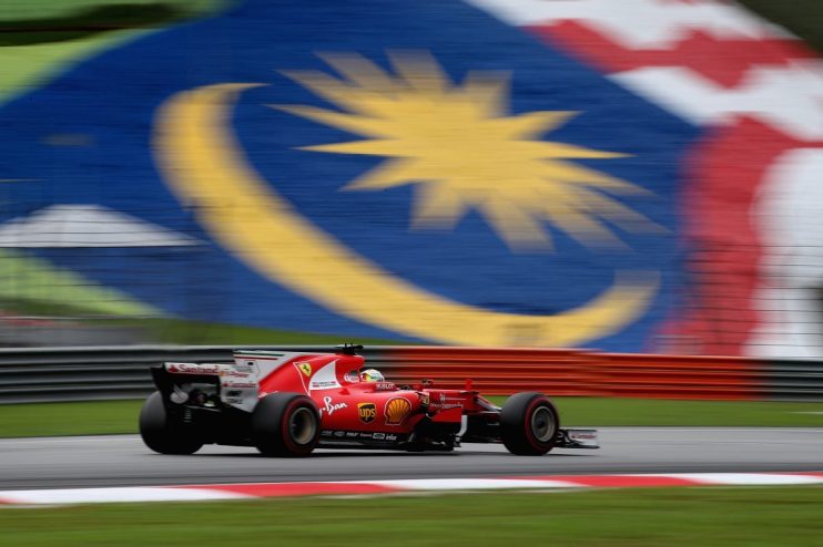 Red Ferrari F1 car speeding past a blurred Malaysian flag banner on a race track