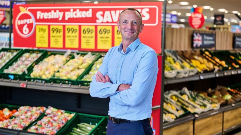 Giles Hurley, Aldi UK CEO, stands in a supermarket produce aisle with fresh fruits and vegetables.
