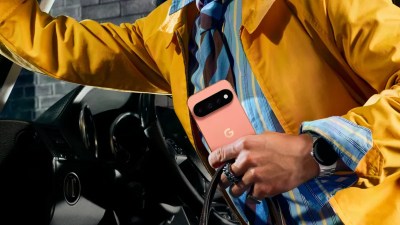 Person in yellow jacket holding a coral Google Pixel phone, with a smartwatch on their wrist, next to a car.