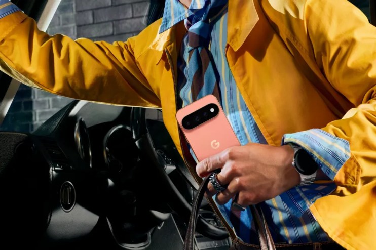 Person in yellow jacket holding a coral Google Pixel phone, with a smartwatch on their wrist, next to a car.