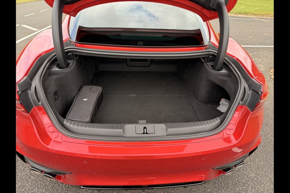 Open trunk of a red sports car, revealing a spacious, carpeted cargo area with a black bag on the left