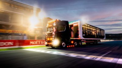 Black Formula 1 hospitality truck driving on a race track at dusk, bright lights and sponsor banners visible.