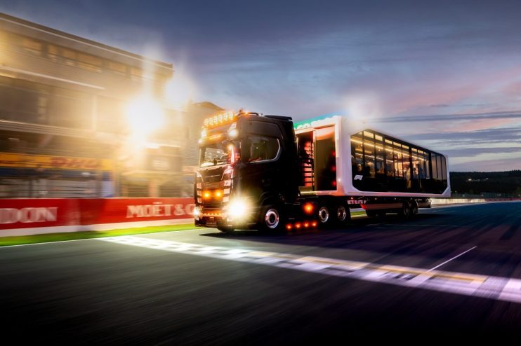 Black Formula 1 hospitality truck driving on a race track at dusk, bright lights and sponsor banners visible.