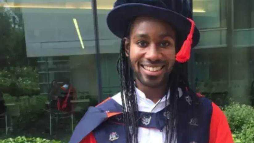 Jason Arday smiling, wearing academic regalia with a blue cap and gown with red accents.