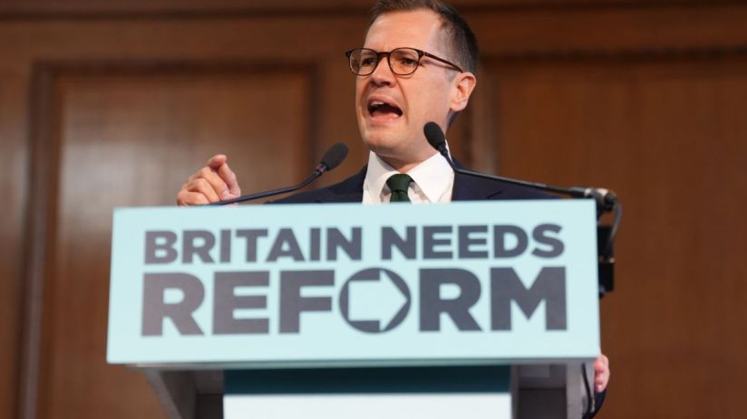 Robert Jenrick speaking at a podium with BRITAIN NEEDS REFORM sign, delivering a speech.