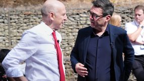 John Healey, in a red tie, speaking with Andy Burnham, wearing glasses and a dark blue jacket, outdoors.