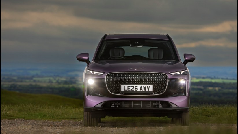 Front view of a purple SUV, possibly a Lepas L8, with headlights on, on a dirt road against a cloudy sky.