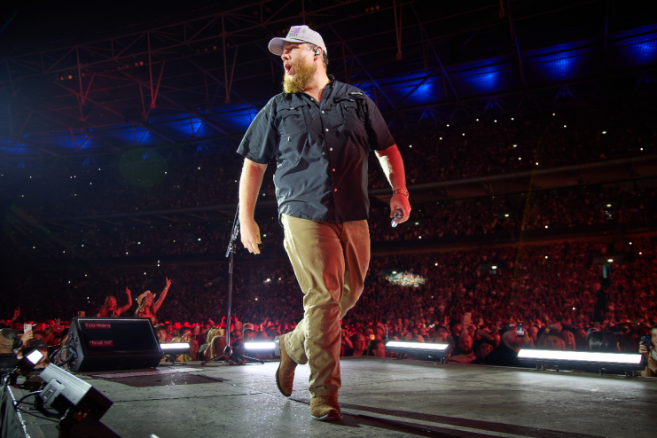 Luke Combs performing on stage at Wembley Stadium, facing right with a microphone, large crowd visible