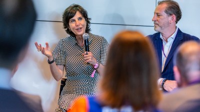 A woman in a polka dot dress speaks into a microphone at a business event, a man in a suit beside her.
