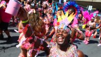 Joyful reveler in vibrant feathered and jeweled costume at Notting Hill Carnival 2025, holding a pink mug.