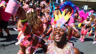 Joyful reveler in vibrant feathered and jeweled costume at Notting Hill Carnival 2025, holding a pink mug.