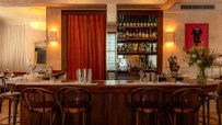 Elegant bar interior with wooden counter, high stools, and shelves of liquor bottles, designed by Justin De Souza.