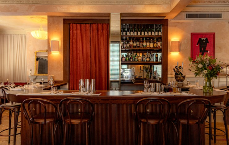 Elegant bar interior with wooden counter, high stools, and shelves of liquor bottles, designed by Justin De Souza.