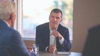Paul Cooper, a man in a navy suit, gestures during a business meeting at a conference table.