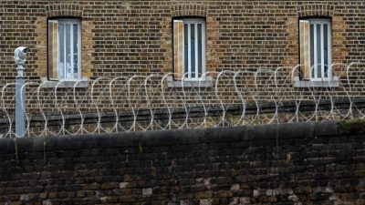 Brick prison wall with barred windows, razor wire, and a security camera.