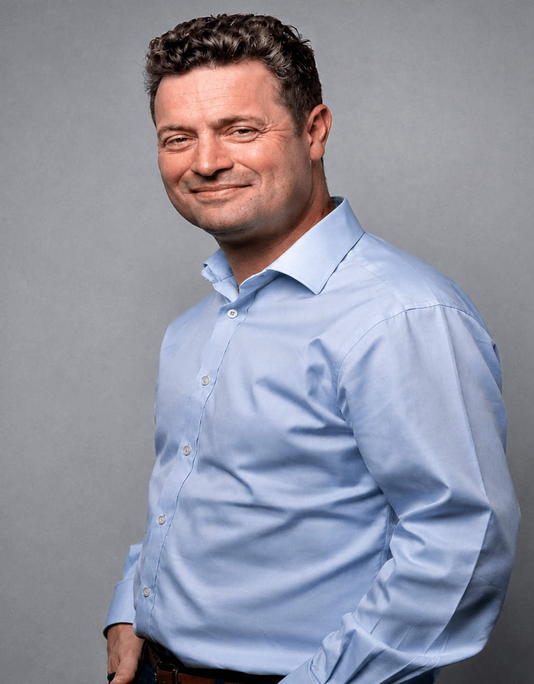 Olly Purnell, a man with short curly brown hair, smiling in a light blue dress shirt, hands in pockets.