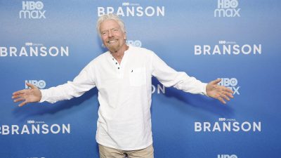 Richard Branson smiling with arms outstretched in front of a blue HBO Max Branson backdrop