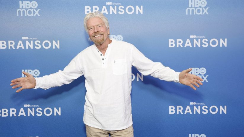 Richard Branson smiling with arms outstretched in front of a blue HBO Max Branson backdrop