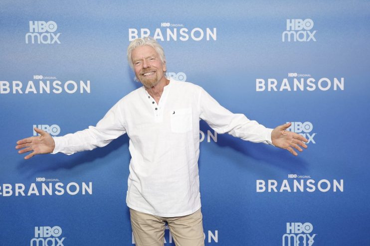 Richard Branson smiling with arms outstretched in front of a blue HBO Max Branson backdrop