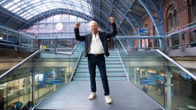 Richard Branson with arms raised in victory on a modern staircase inside St. Pancras International Station