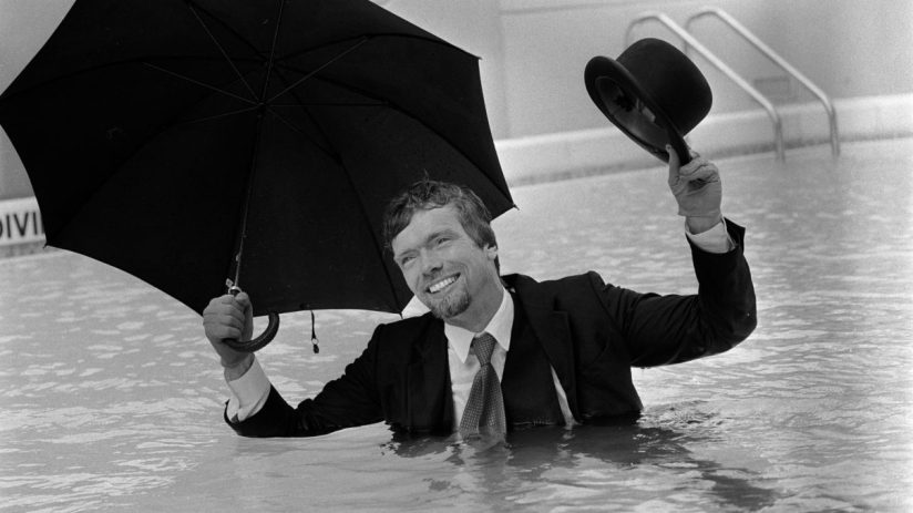 Richard Branson in a suit, holding an umbrella and bowler hat, smiling in a swimming pool