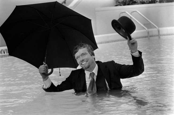 Richard Branson in a suit, holding an umbrella and bowler hat, smiling in a swimming pool