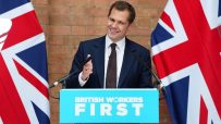 Robert Jenrick smiling at a podium with BRITISH WORKERS FIRST text, flanked by Union Jack flags at a press conference.