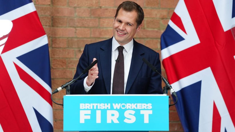 Robert Jenrick smiling at a podium with BRITISH WORKERS FIRST text, flanked by Union Jack flags at a press conference.