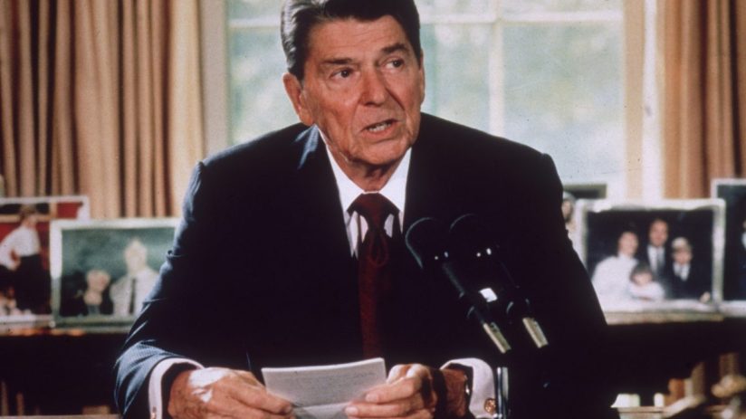 Ronald Reagan speaking at a podium with microphones, holding a paper, family photos visible in the background