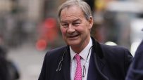 Rupert Lowe, former Southampton FC chairman, smiles while holding files on a city street, wearing a suit and pink tie