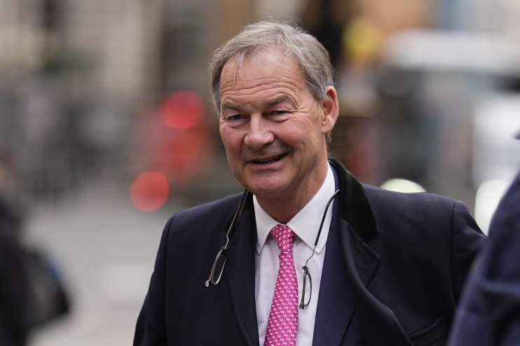 Rupert Lowe, former Southampton FC chairman, smiles while holding files on a city street, wearing a suit and pink tie