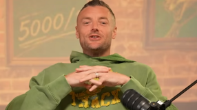 Man in a green hoodie speaking into a microphone, with a 5000 sign in the background.