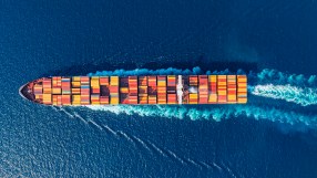 Aerial view of a large container ship moving through deep blue ocean waters, leaving a white wake.
