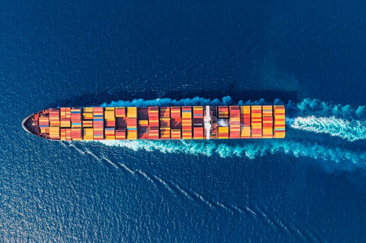 Aerial view of a large container ship moving through deep blue ocean waters, leaving a white wake.