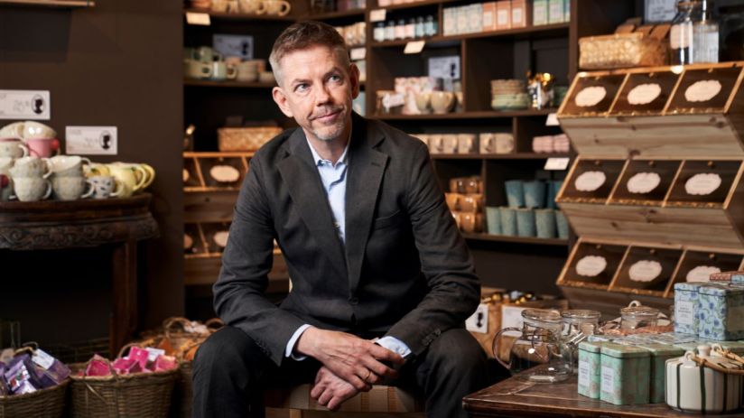 Man in suit seated in a Sostrene Grene store, surrounded by homeware and gift items.