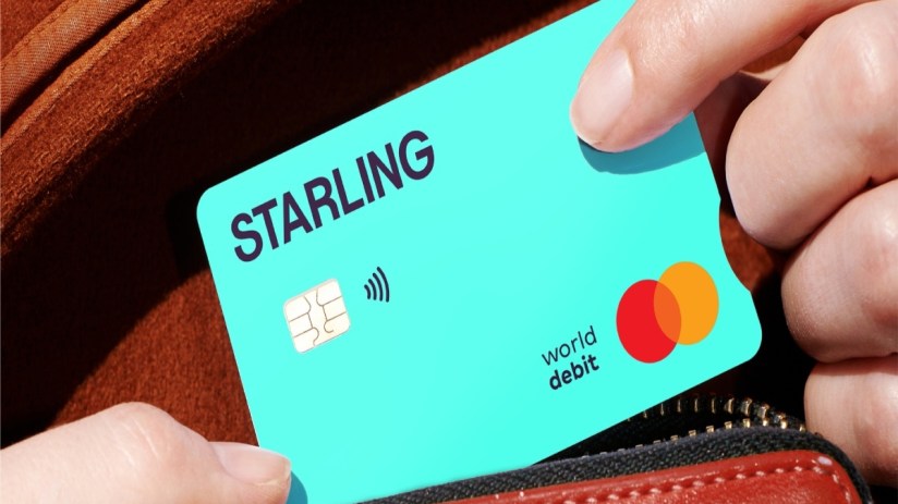 Hand inserting a turquoise Starling Bank PCA debit card with Mastercard logo into a brown wallet.