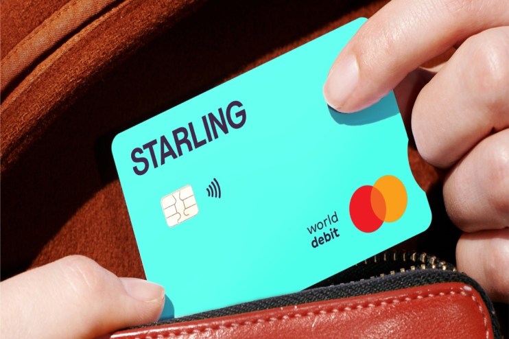 Hand inserting a turquoise Starling Bank PCA debit card with Mastercard logo into a brown wallet.