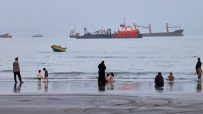 People on a beach with cargo ships and a small boat in the Strait of Hormuz