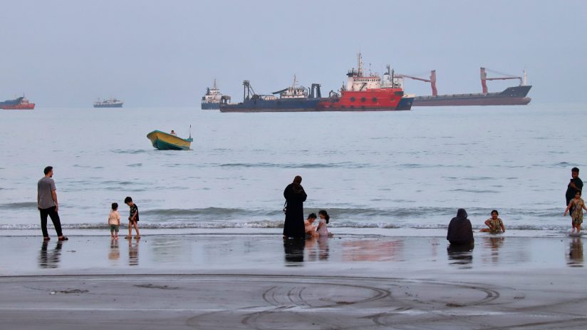 People on a beach with cargo ships and a small boat in the Strait of Hormuz