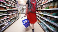 Shopper with red backpack and blue basket walking through a supermarket aisle filled with groceries