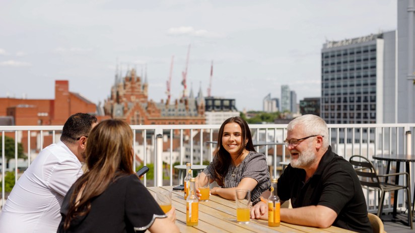 Four colleagues on a sunny rooftop terrace, enjoying drinks and conversation with city buildings in the background.