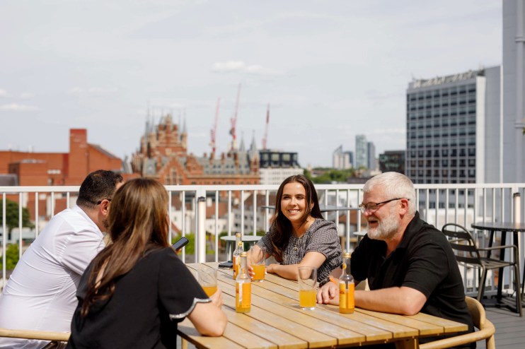 Four colleagues on a sunny rooftop terrace, enjoying drinks and conversation with city buildings in the background.