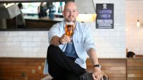 Smiling man in light blue shirt holding up a glass of amber beer in a bar setting