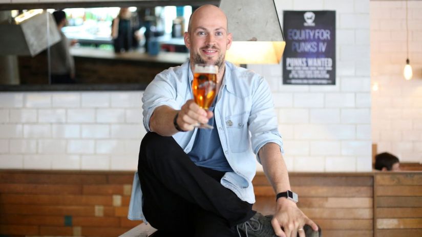 Smiling man in light blue shirt holding up a glass of amber beer in a bar setting