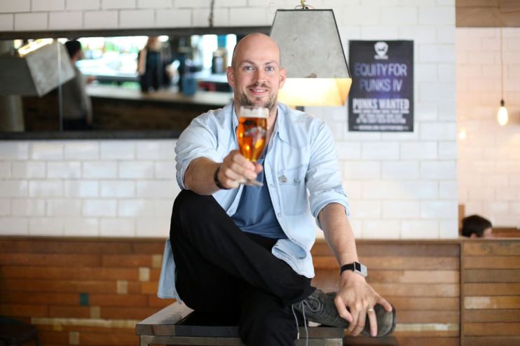 Smiling man in light blue shirt holding up a glass of amber beer in a bar setting