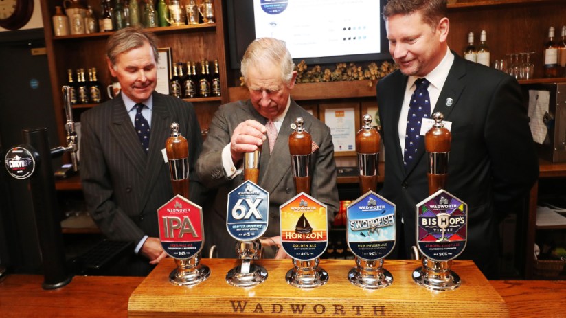 King Charles III pulls a pint at Wadworth Brewery with brewery staff, showcasing beer taps.